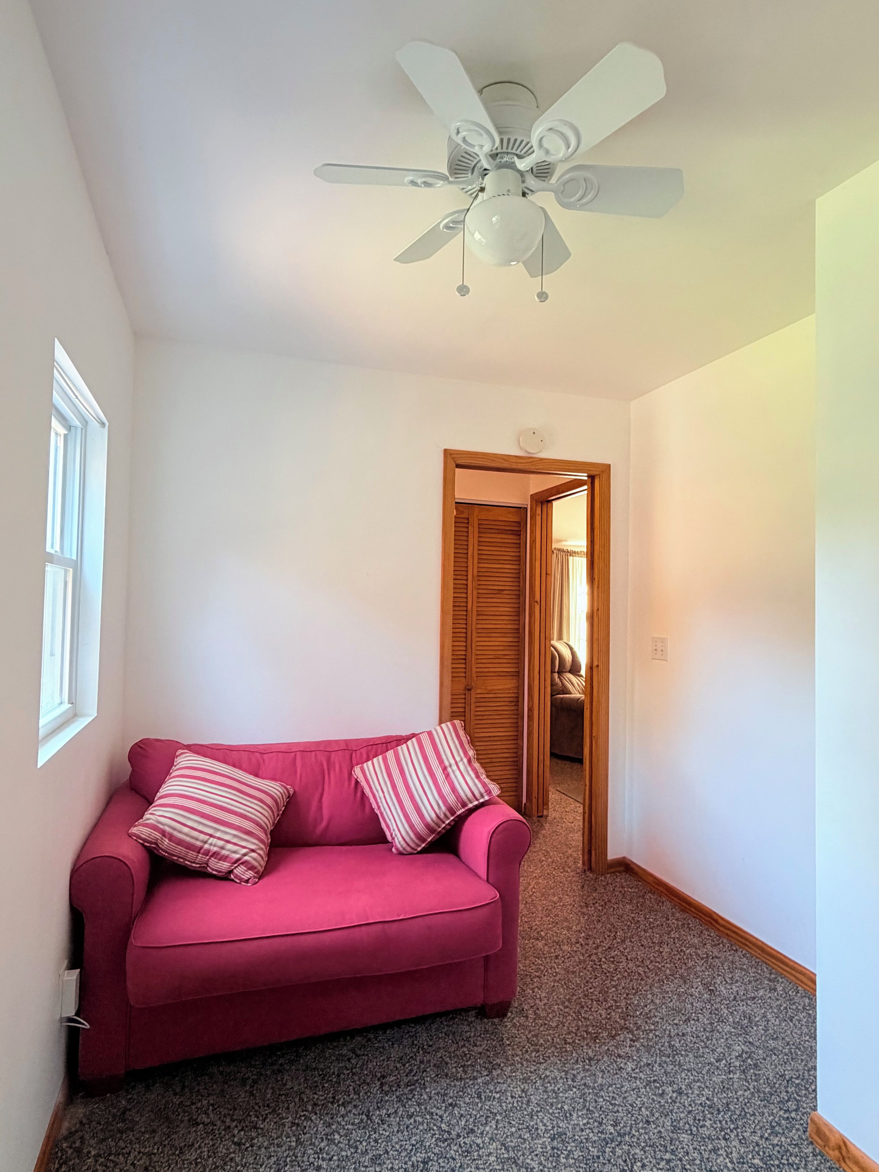Una habitación con un sofá rojo, un ventilador de techo, una puerta de madera y suelo alfombrado.