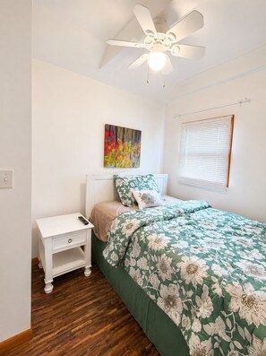A bedroom with a bed, nightstand, ceiling fan, and a window with blinds.