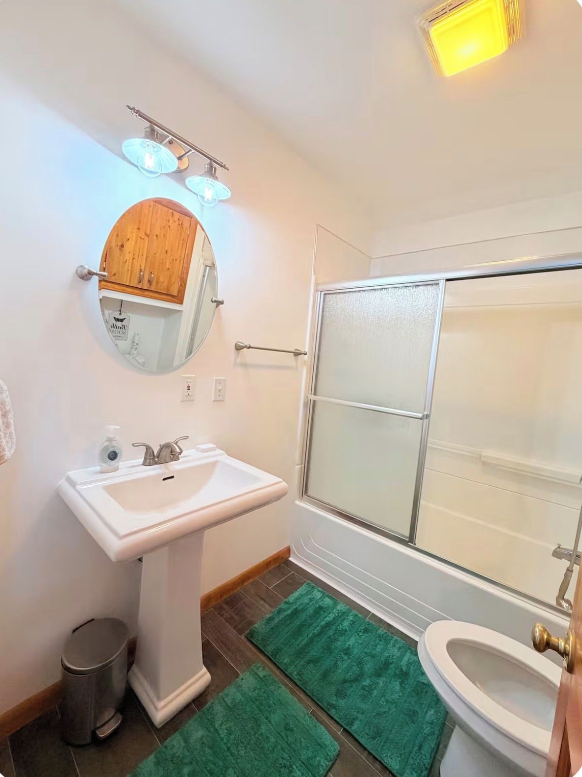 Ein Badezimmer mit einem Standwaschbecken, einem Spiegel, einem Holzkabinet und einer Duschkabine, sowie grünen Teppichen.