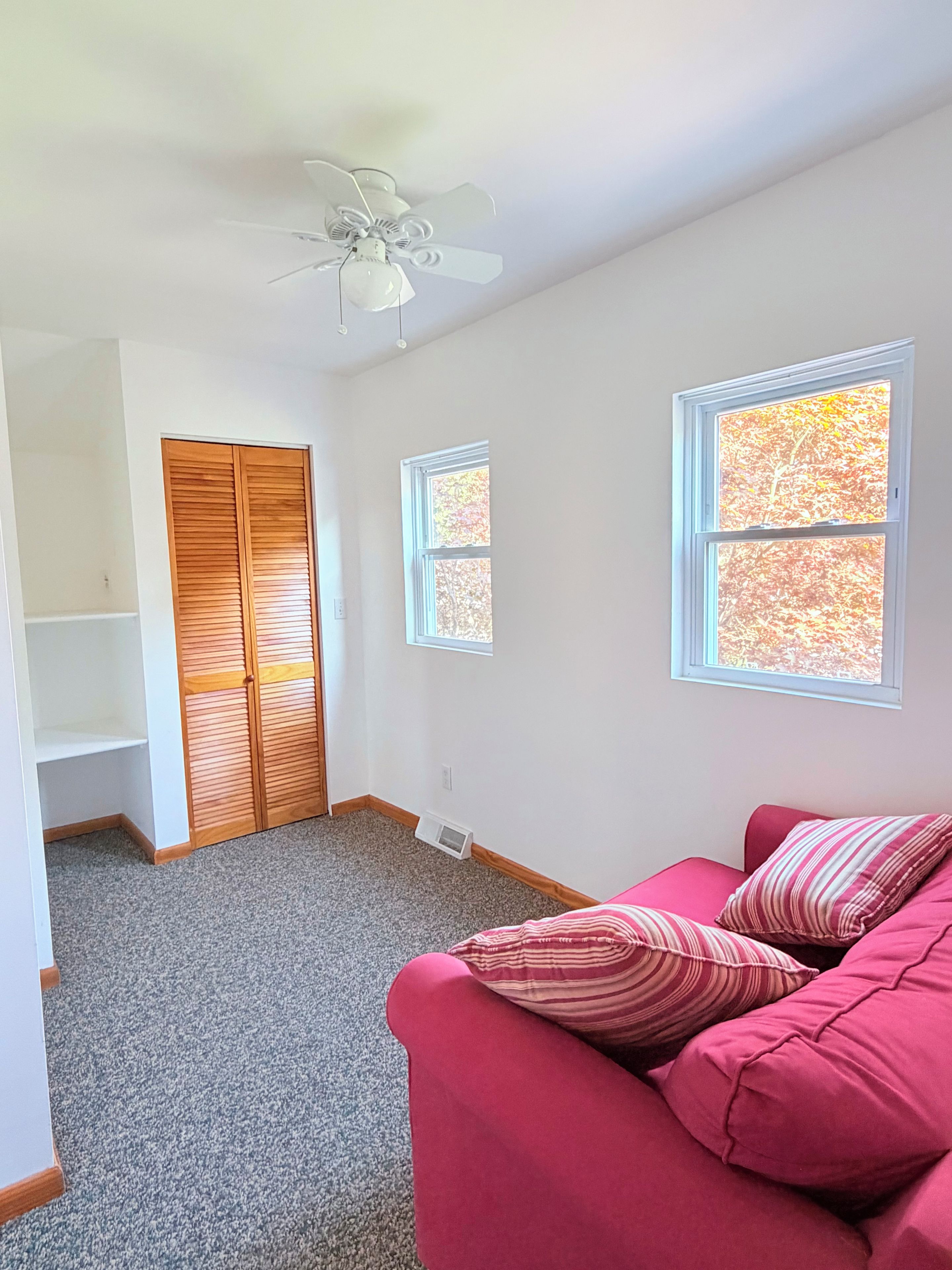 Ein Zimmer mit Deckenventilator, Schrank, Fenster mit Herbstlaub und einem roten Sofa.
