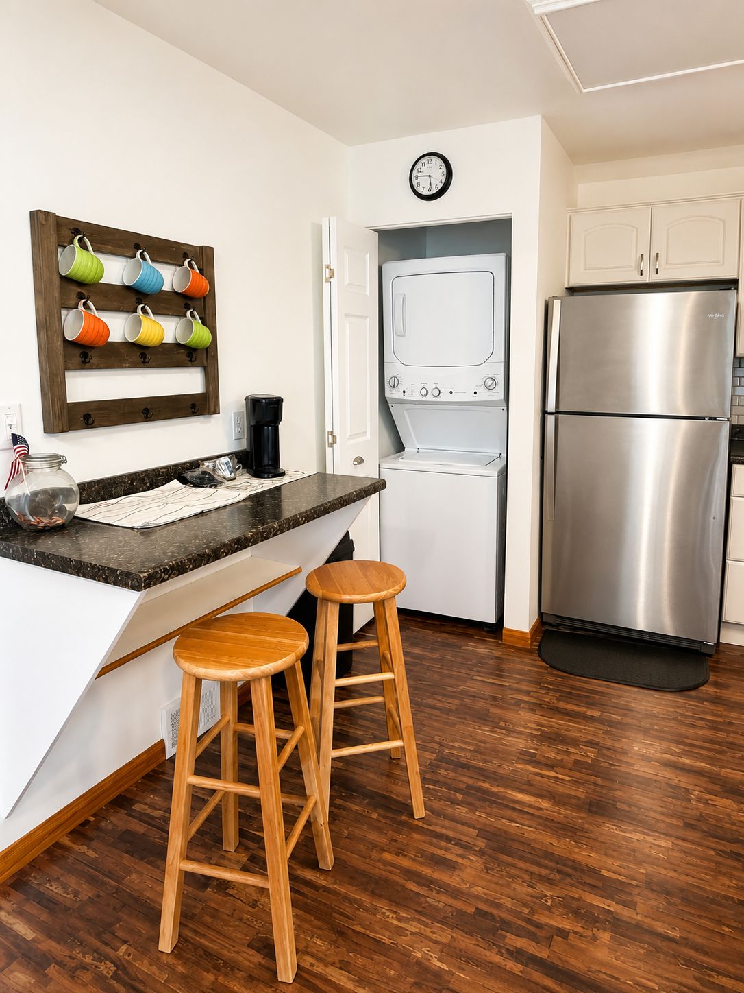 A kitchen with a stainless steel refrigerator, a washing machine, wooden stools, a coffee maker, and a clock on the wall.