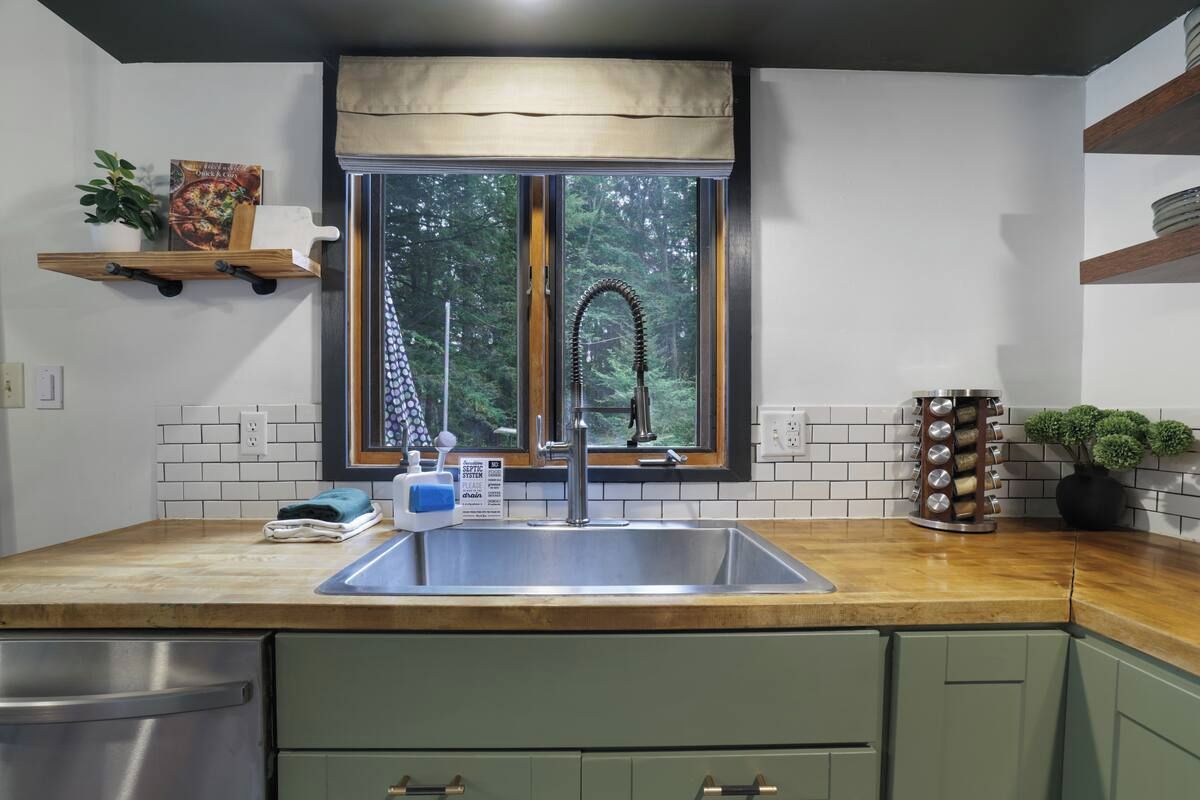 Een keuken met een RVS spoelbak, een raam met uitzicht op bomen, en een houten aanrecht.