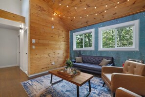 Ein gemütliches Zimmer mit Holzbalkendecke, zwei Fenstern mit Blick auf Bäume, einem Sofa, einem Couchtisch und einem Teppich.