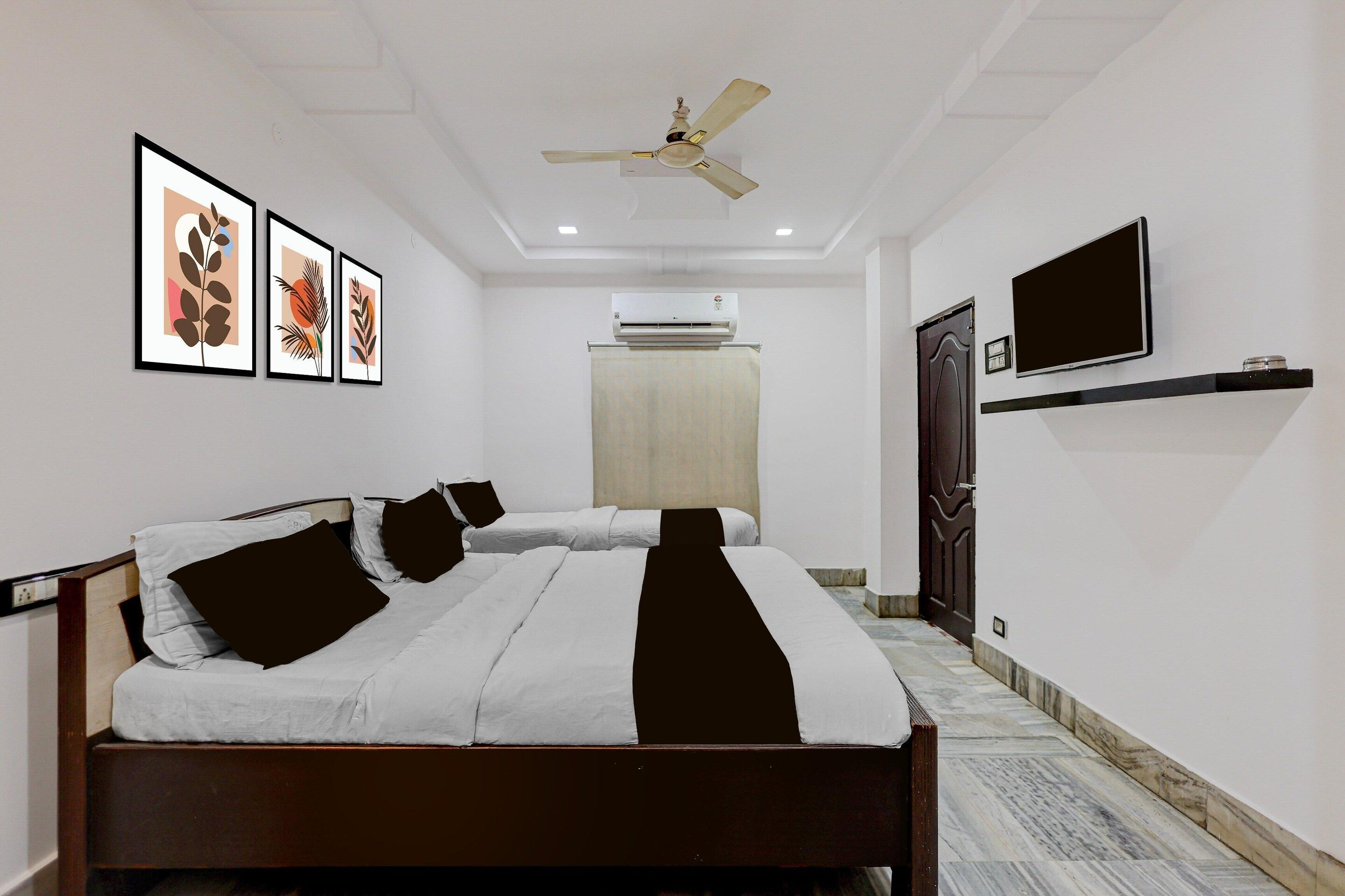 Une chambre d’hôtel équipée d’un lit, d’un ventilateur de plafond, d’un climatiseur, d’un téléviseur à écran plat et d’œuvres d’art encadrées accrochées au mur.