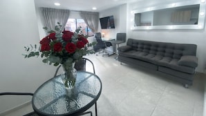 Uma sala de estar moderna com um sofá cinza, uma mesa de vidro com um vaso de flores vermelhas, uma TV na parede e uma janela com cortinas.