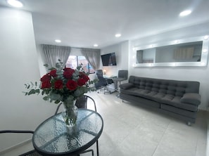A modern living room with a grey sofa, a glass table with a vase of red flowers, a TV on the wall, and a window with curtains. - New modern studio apartment in Dosquebradas, Pereira (Dosquebradas)
