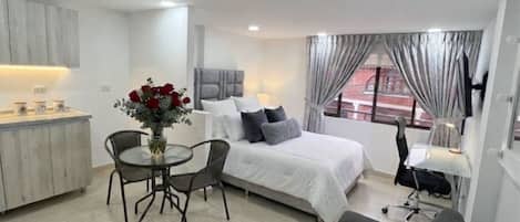 Habitación de hotel moderna con cama, una mesa de comedor con sillas, una pequeña cocina y una ventana con cortinas.
