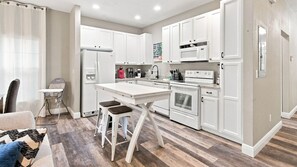 A modern kitchen with white cabinetry, a central island with stools, and a dining area with a sofa and a patterned throw pillow. - Coral Cay 4BR Townhouse Near Disney & Universal (Kissimmee)