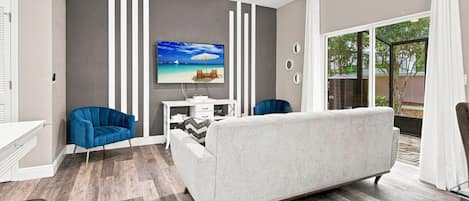 A modern living room with a grey sofa, blue chairs, a white desk, and a TV mounted on the wall.