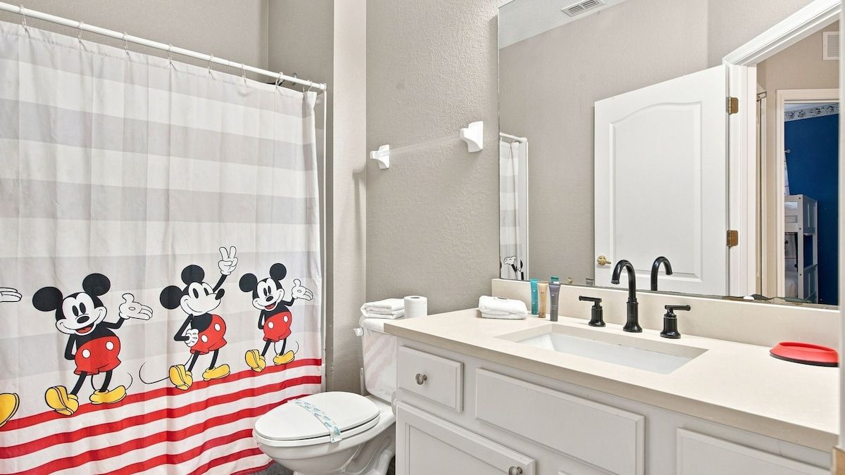 Ein Badezimmer mit Mickey-Mouse-Duschvorhang, Toilette in Weiß, Waschbecken mit Spiegel und Handtuchhalter.