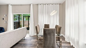 A modern living room with a sofa, a glass-top coffee table, and four chairs. There is a sliding glass door leading to an outdoor area. - Coral Cay 4BR Townhouse Near Disney & Universal (Kissimmee)