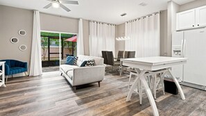 A modern living room with a sofa, dining table, and chairs, a ceiling fan, and a large window with curtains. - Coral Cay 4BR Townhouse Near Disney & Universal (Kissimmee)