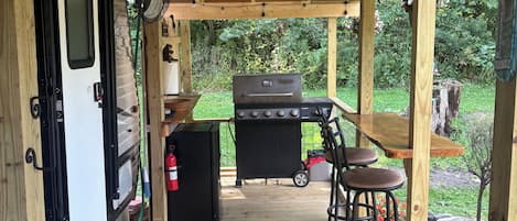 A covered outdoor area with a grill, seating, and a door leading to a vehicle.