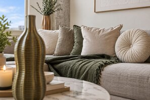 Ein gemütliches Wohnzimmer mit einer Couch, Kissen, einer grünen Decke, einer Vase und einer Kerze auf einem Tisch.