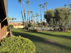 A well-maintained lawn with a variety of trees, including palm trees, and a brick building with a garage. - Amazing Chaparral Country Club Updated Town Home on 6th Tee Box. (Palm Desert)