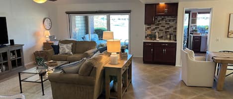 A living room with a sofa, coffee table, and a television. There is a kitchen area with cabinets and a window.