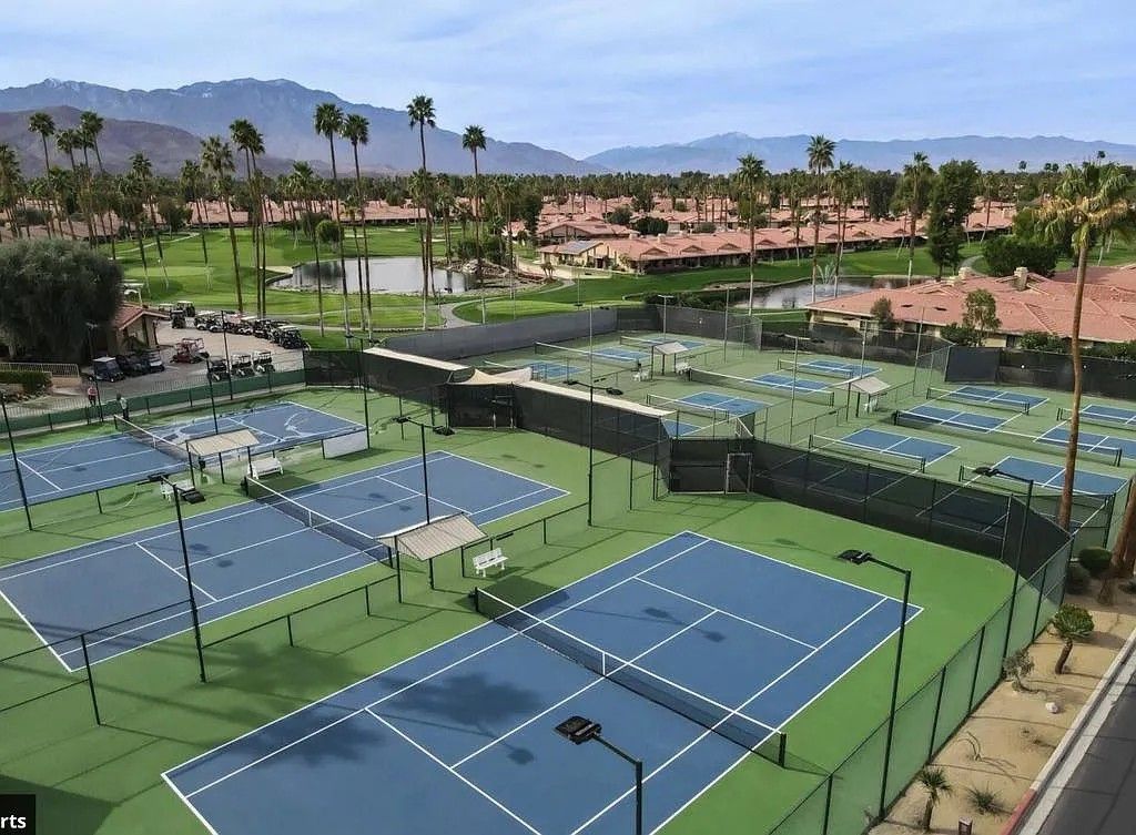 Ein Tennisplatzkomplex mit mehreren Plätzen, einer überdachten Zuschauertribüne und Blick auf eine Wohngegend mit Palmen und Bergen im Hintergrund.