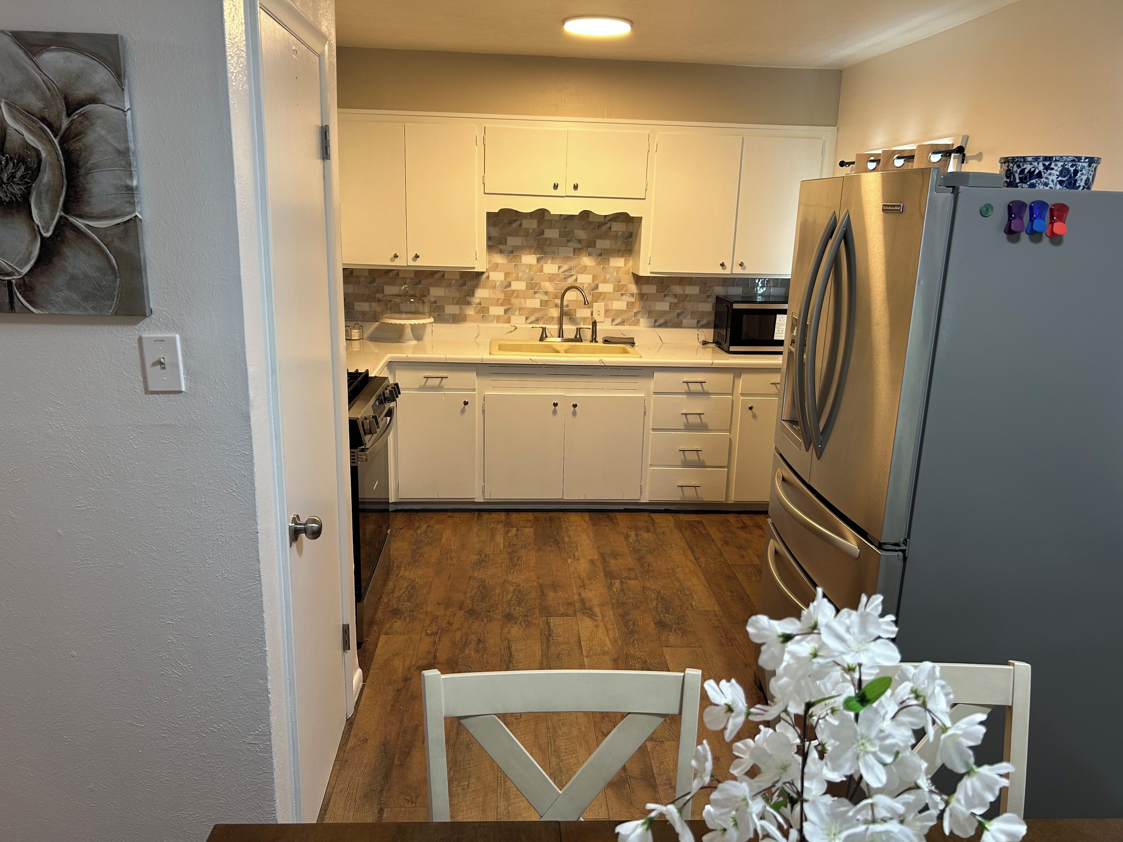 Una cocina con armarios blancos, un refrigerador de acero inoxidable y piso de madera.