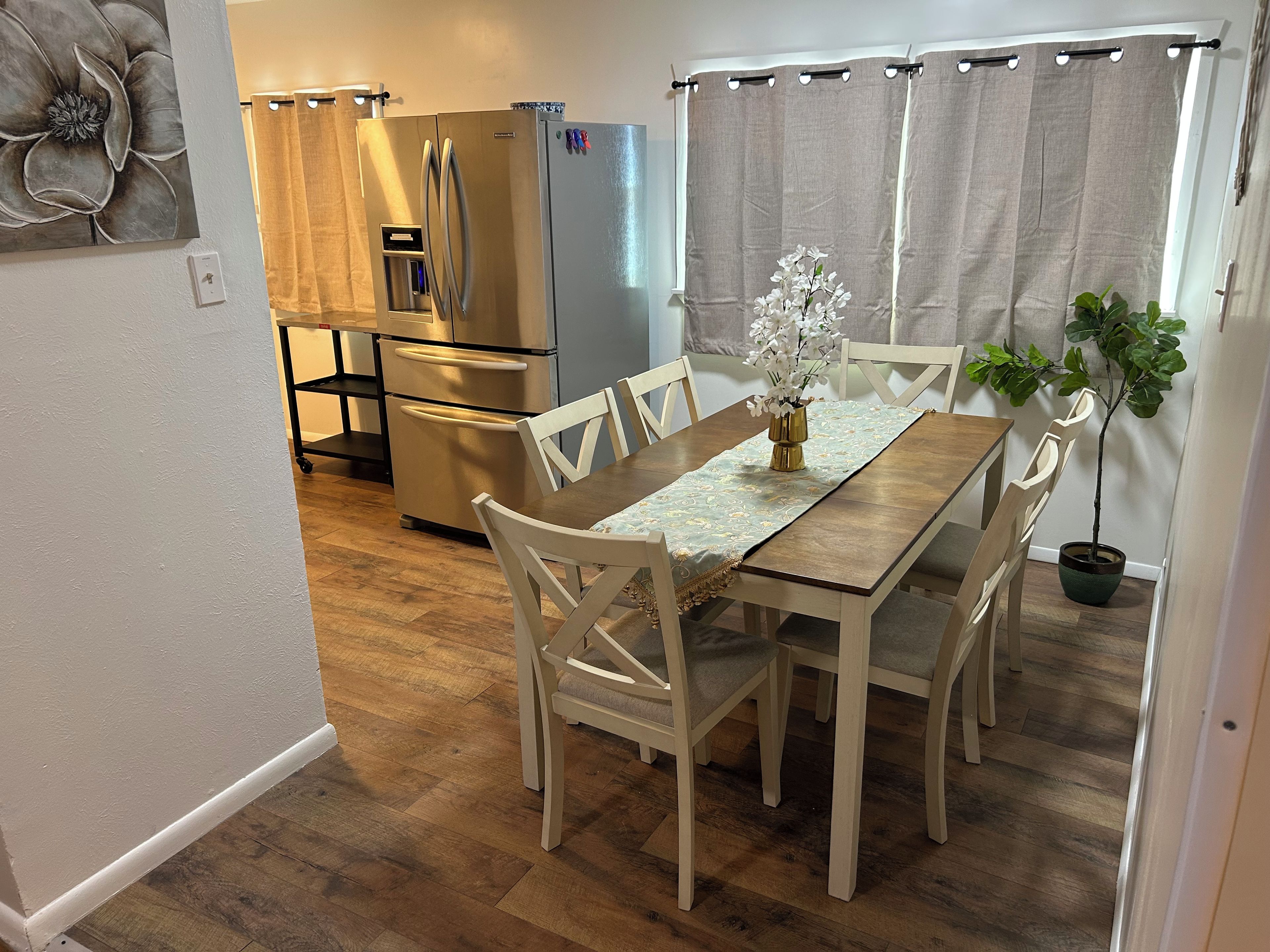 A dining area with a wooden table, white chairs, a vase with flowers, and a refrigerator in the background.