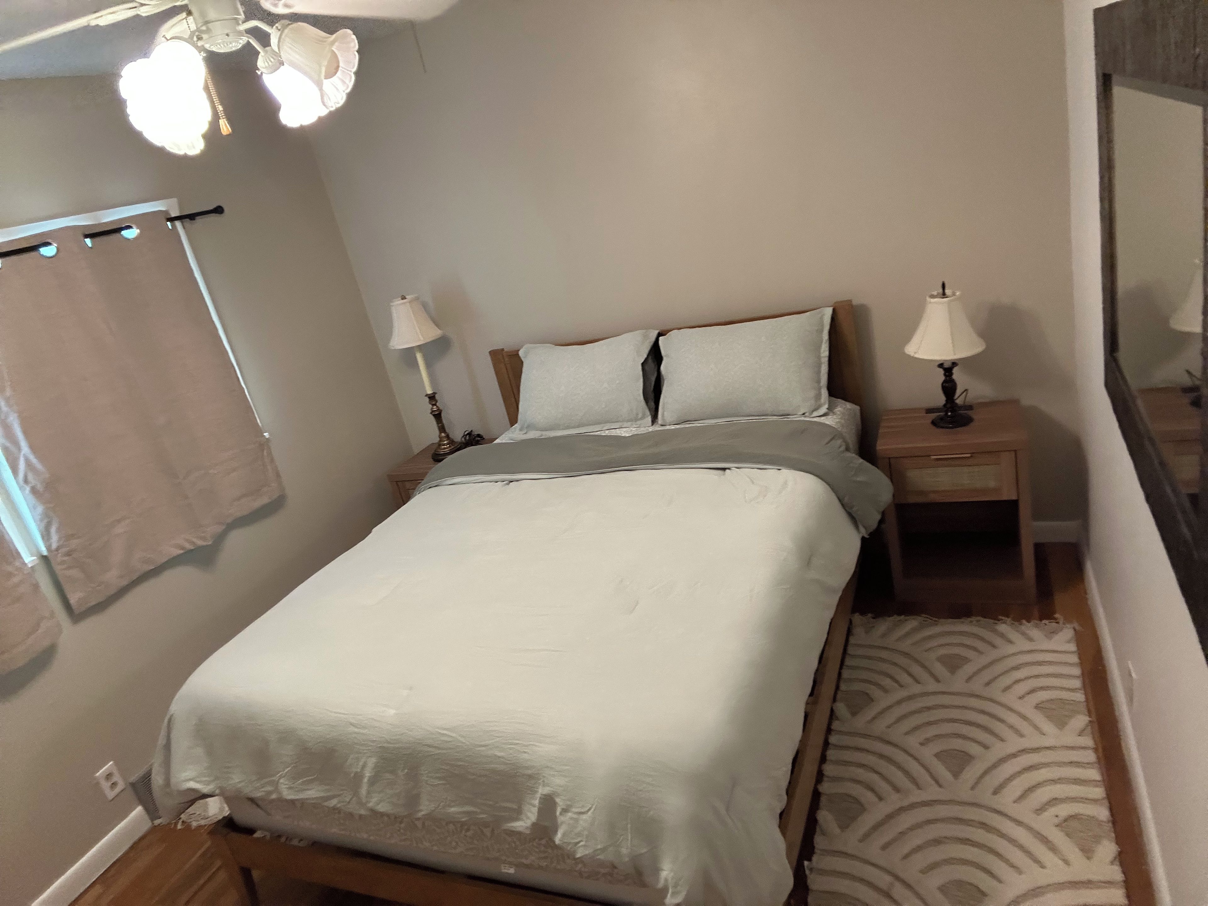 A bedroom with a bed, two bedside tables with lamps, a ceiling light fixture, and a patterned rug.