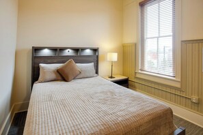 Ein Hotelzimmer mit Bett, Nachttisch mit Lampe, Fenster mit Jalousien und Heizkörper.