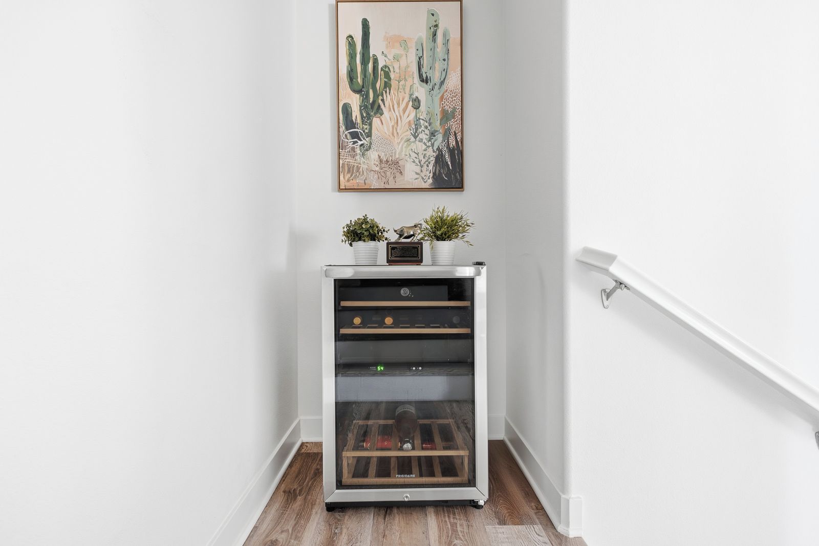 A wine refrigerator with a digital display, a cactus painting above it, and a white railing to the right.