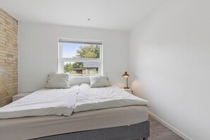 Ein Schlafzimmer mit einem Bett, einem Nachttisch, einer Lampe und einem Fenster mit Blick auf ein Haus und Bäume.