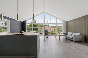 Eine moderne Küche mit einer großen Kochinsel, einem Essbereich inklusive Sofa und Tisch, sowie großen Fenstern mit Blick nach draußen.