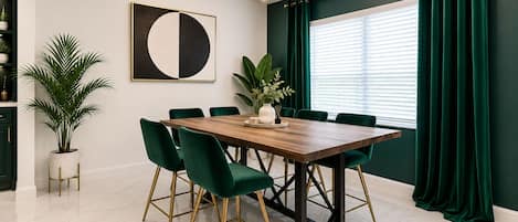 A dining area with a wooden table, green upholstered chairs, a potted plant, and a modern chandelier.