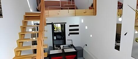Habitación compacta con una escalera de madera que conduce a una zona superior, una kitchenette y un espacio de descanso con sofá.