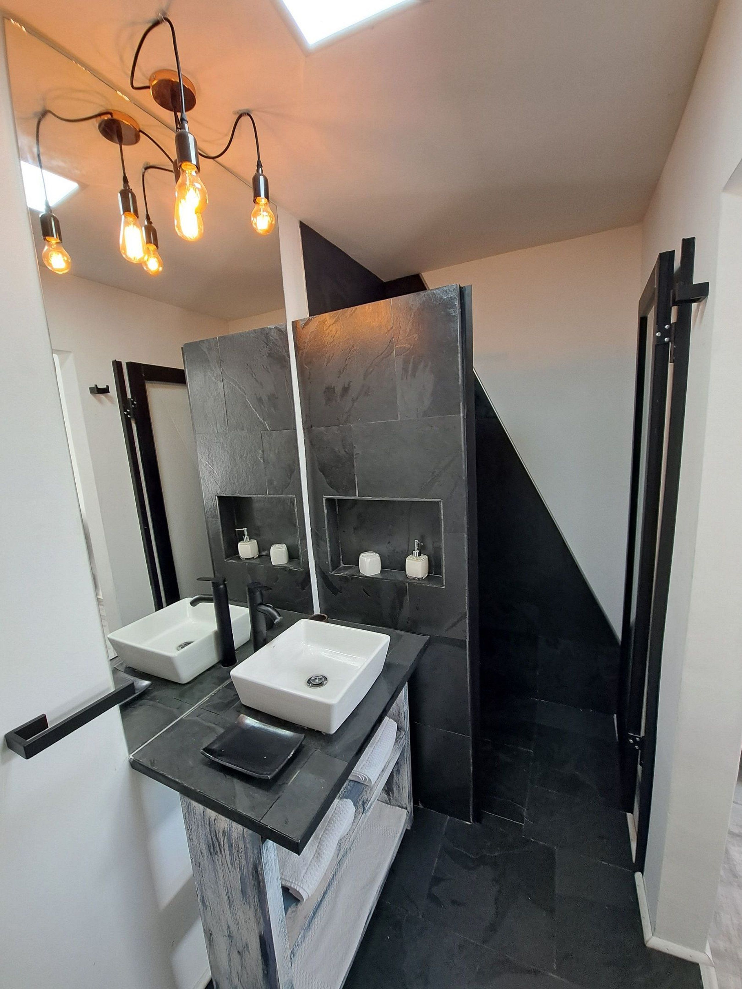 A modern bathroom with two sinks, a black countertop, and a skylight above.