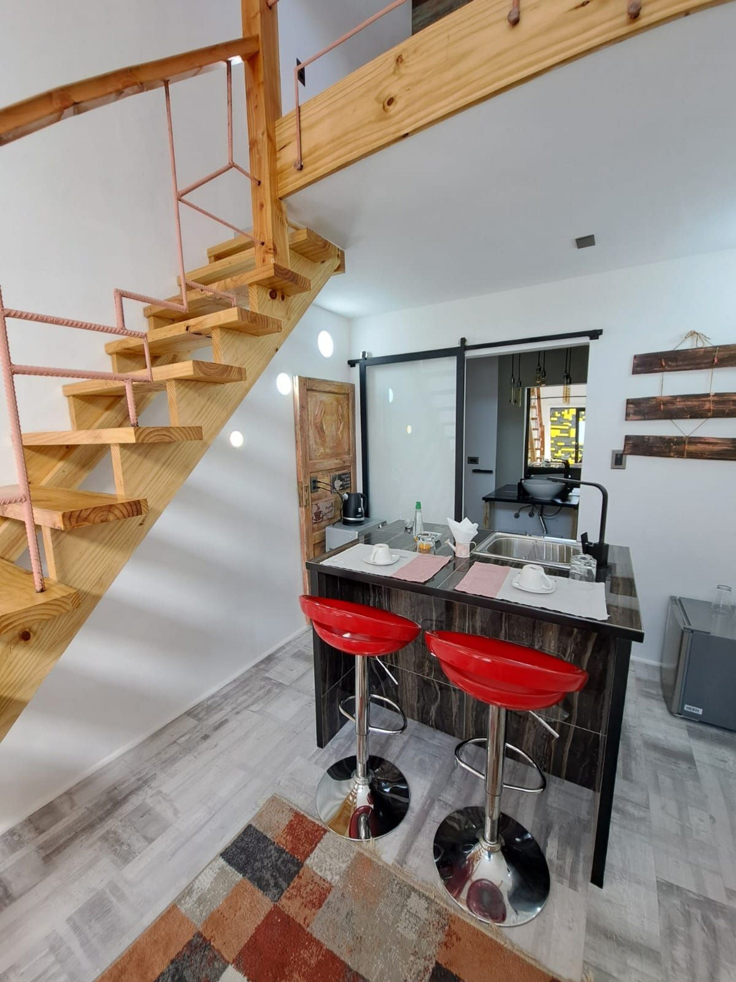 A modern kitchen with a wooden staircase, a central island with red bar stools, and a large mirror above the island.