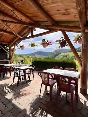 Une terrasse extérieure avec des tables et des chaises, des poutres en bois, des pots de fleurs suspendus et une vue sur des collines verdoyantes.