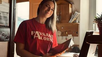 A person in a red shirt standing in a room with a wooden desk, a book, and a potted plant.