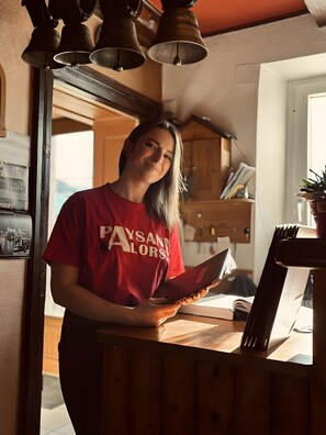 A person in a red shirt standing in a room with a wooden desk, a book, and a potted plant. - Guest Room "Chambre Sophie" with Mountain View and Shared Terrace (Orbey)