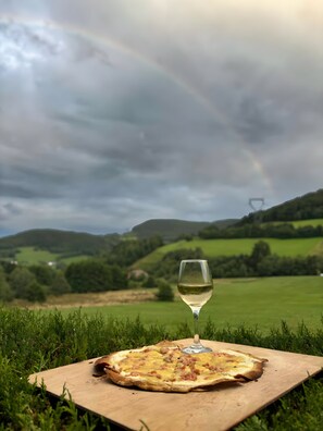 A pizza on a wooden board with a glass of white wine in a grassy field under a cloudy sky.