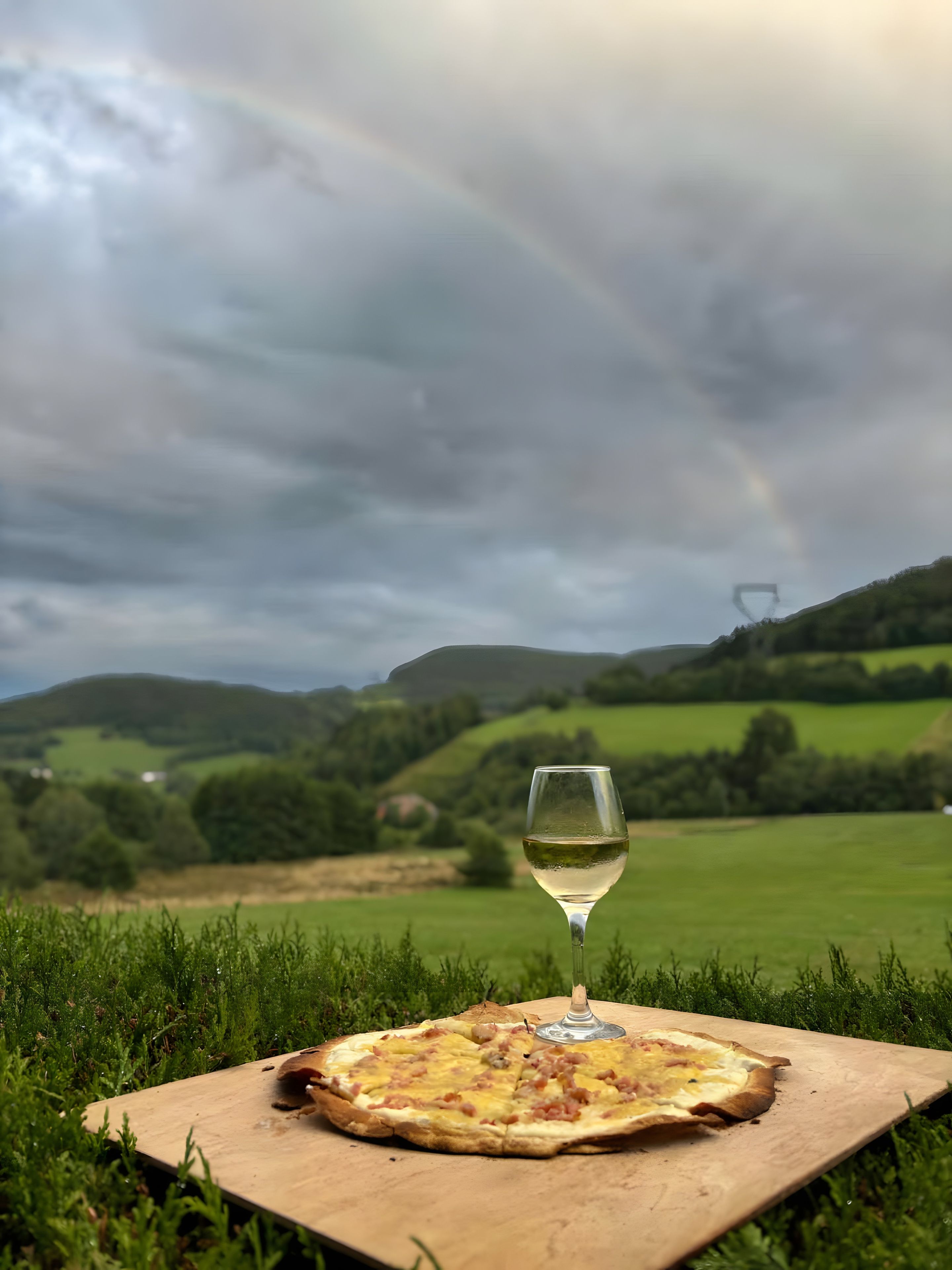 Une pizza servie sur une planche en bois, accompagnée d’un verre de vin blanc, dans un champ verdoyant sous un ciel nuageux.