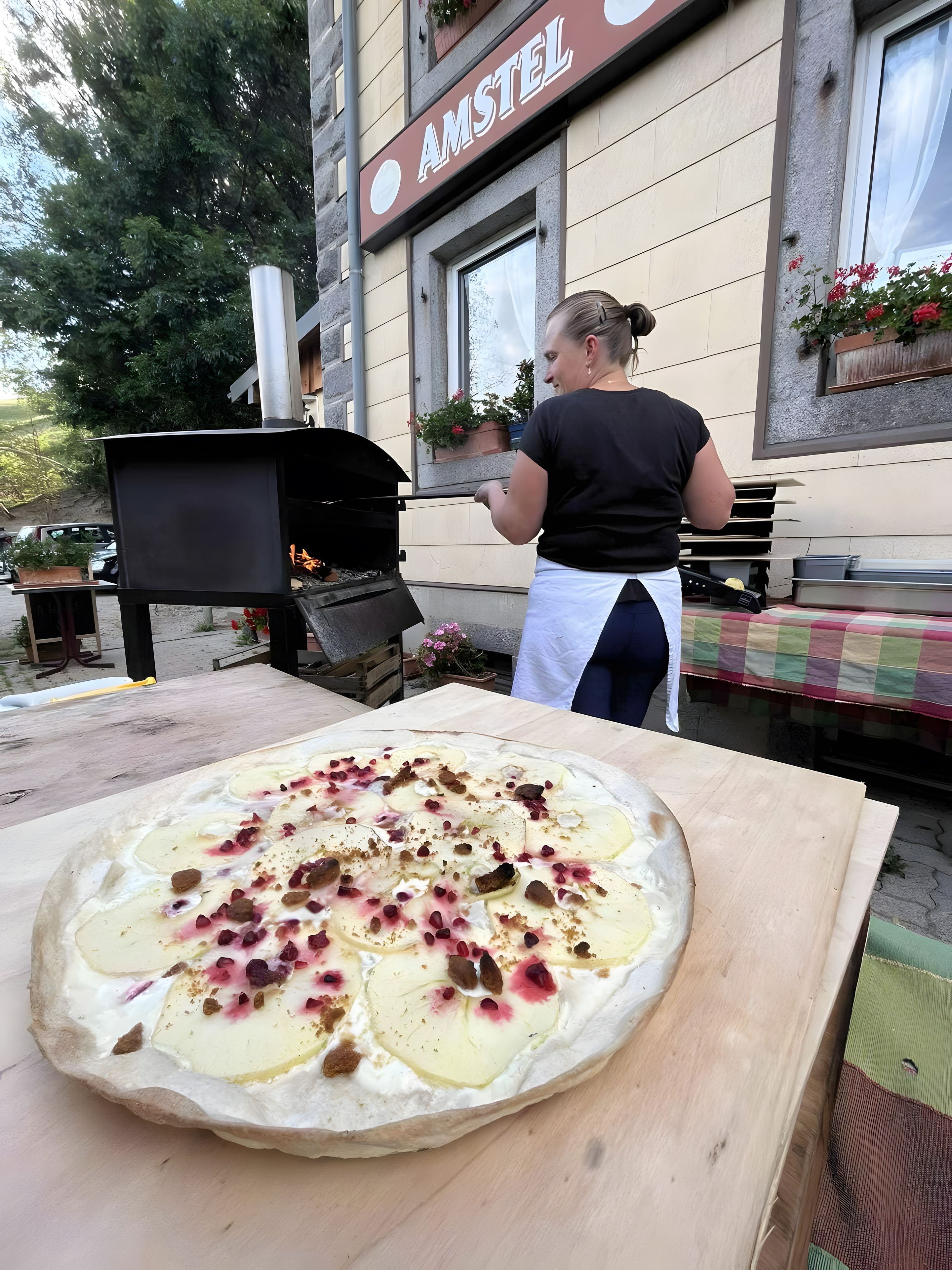 Una pizza sobre una mesa de madera con ingredientes, una mujer con delantal y un edificio con el letrero 'AMSTEL'.