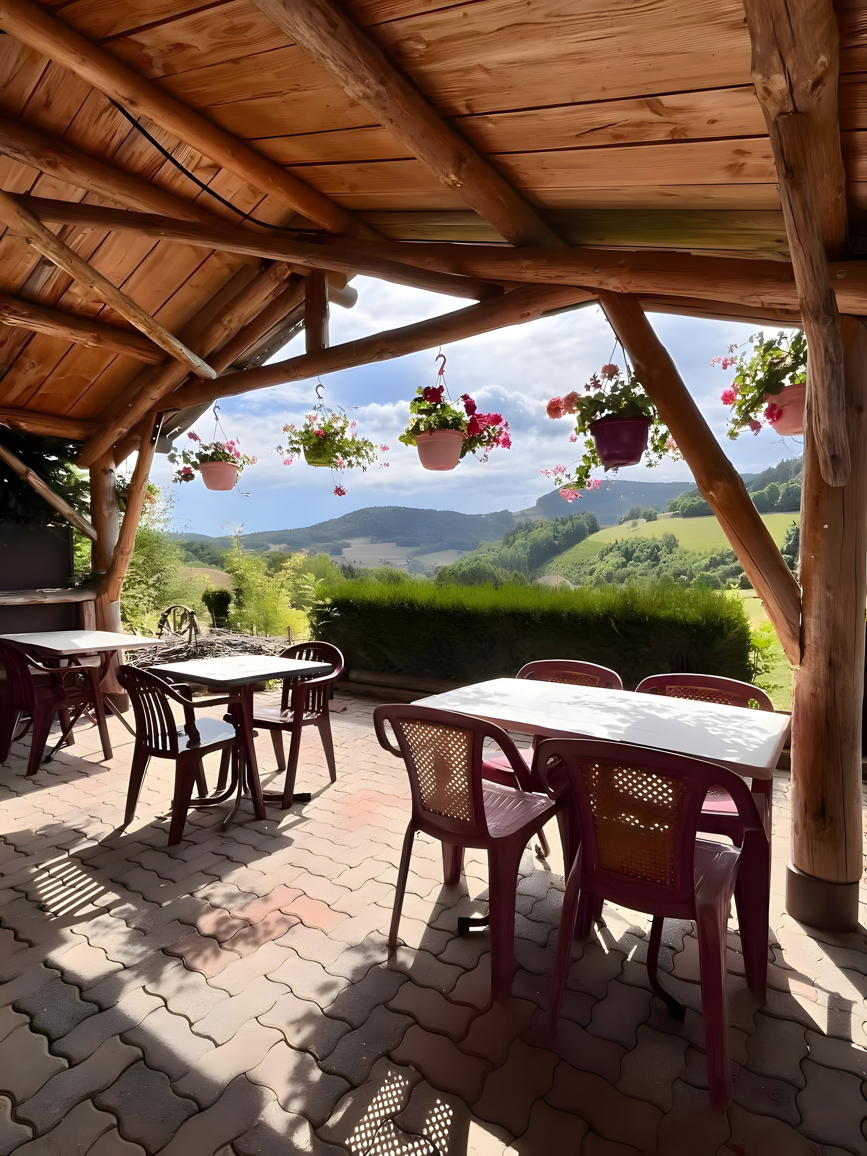 An outdoor patio with tables and chairs, wooden beams, hanging flower pots, and a view of green hills.