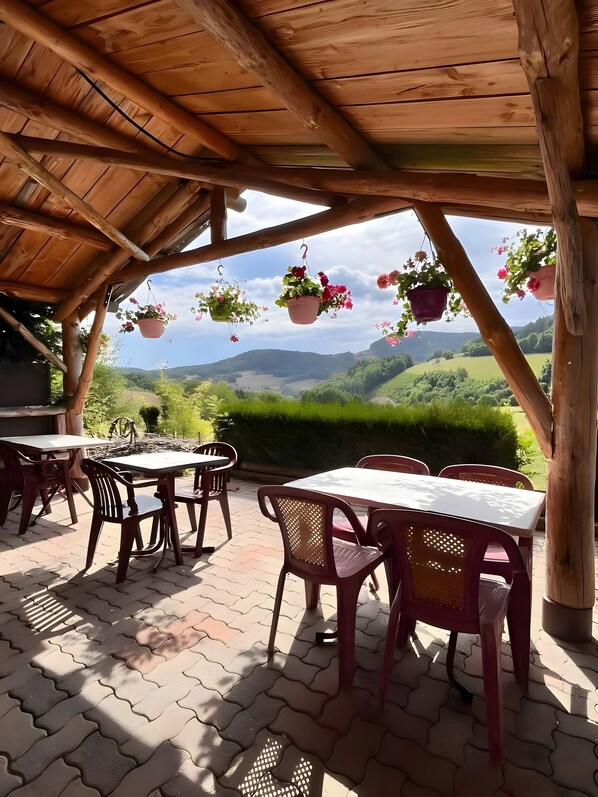 An outdoor patio with tables and chairs, wooden beams, hanging flower pots, and a view of green hills.