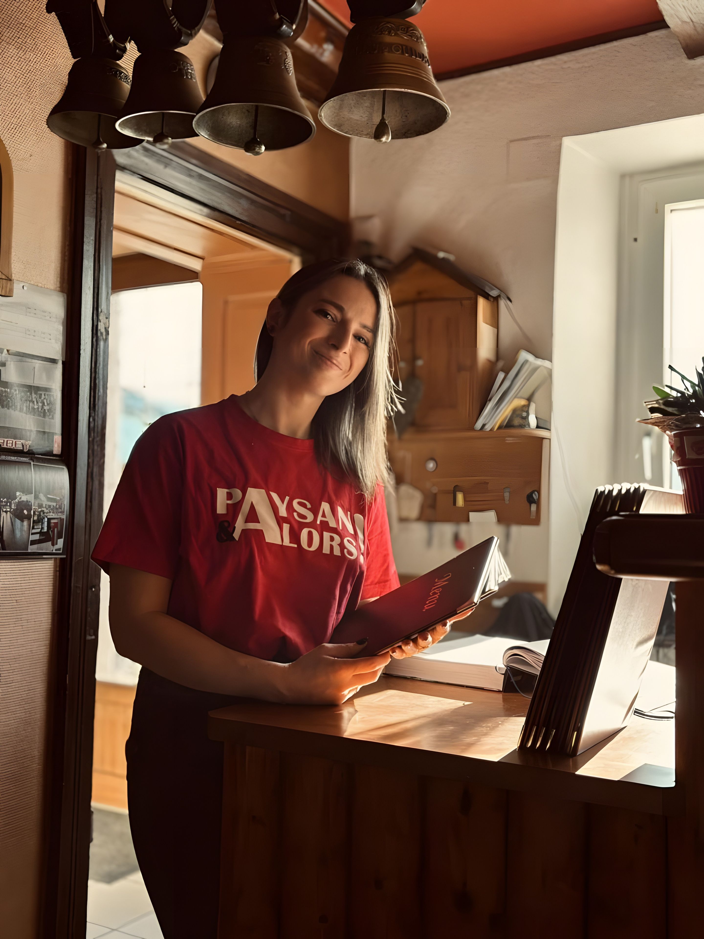 Une personne portant une chemise rouge se tient dans une pièce où se trouvent un bureau en bois, un livre et une plante en pot.