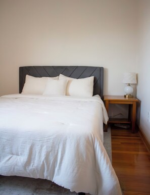 A neatly made bed with a grey headboard, a bedside table with a lamp, and a wooden floor. - Comfortable 3BR getaway 5 min from Midway Airport and 15 min to Downtown. (Chicago)
