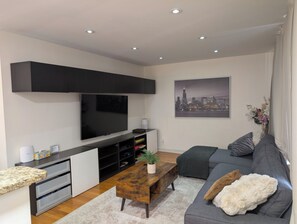 A modern living room with a flat-screen TV, built-in shelving, a grey sofa, and a cityscape painting. - Comfortable 3BR getaway 5 min from Midway Airport and 15 min to Downtown. (Chicago)