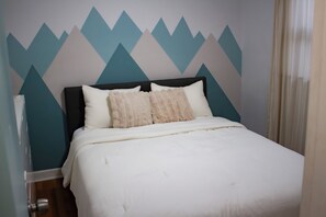 Ein Bett mit weißer Decke, zwei Kissen, einem dunklen Kopfteil und einer Wand mit geometrischem Muster.