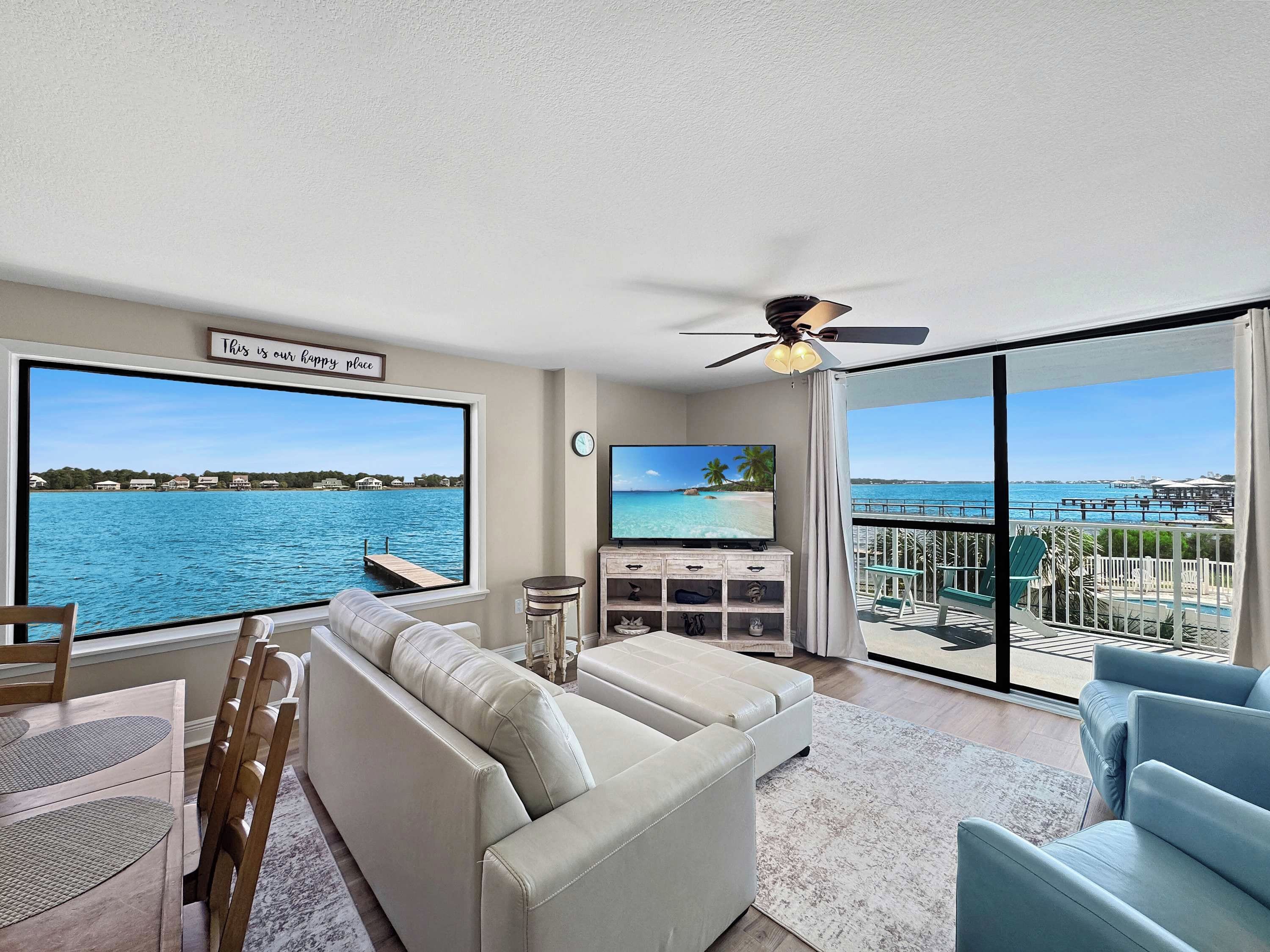 A living room with a large window, a ceiling fan, a flat-screen TV, and a view of the ocean.