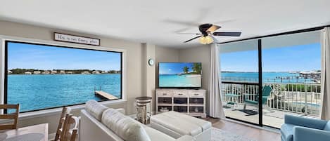 A living room with a large window, a ceiling fan, a flat-screen TV, and a view of the ocean.