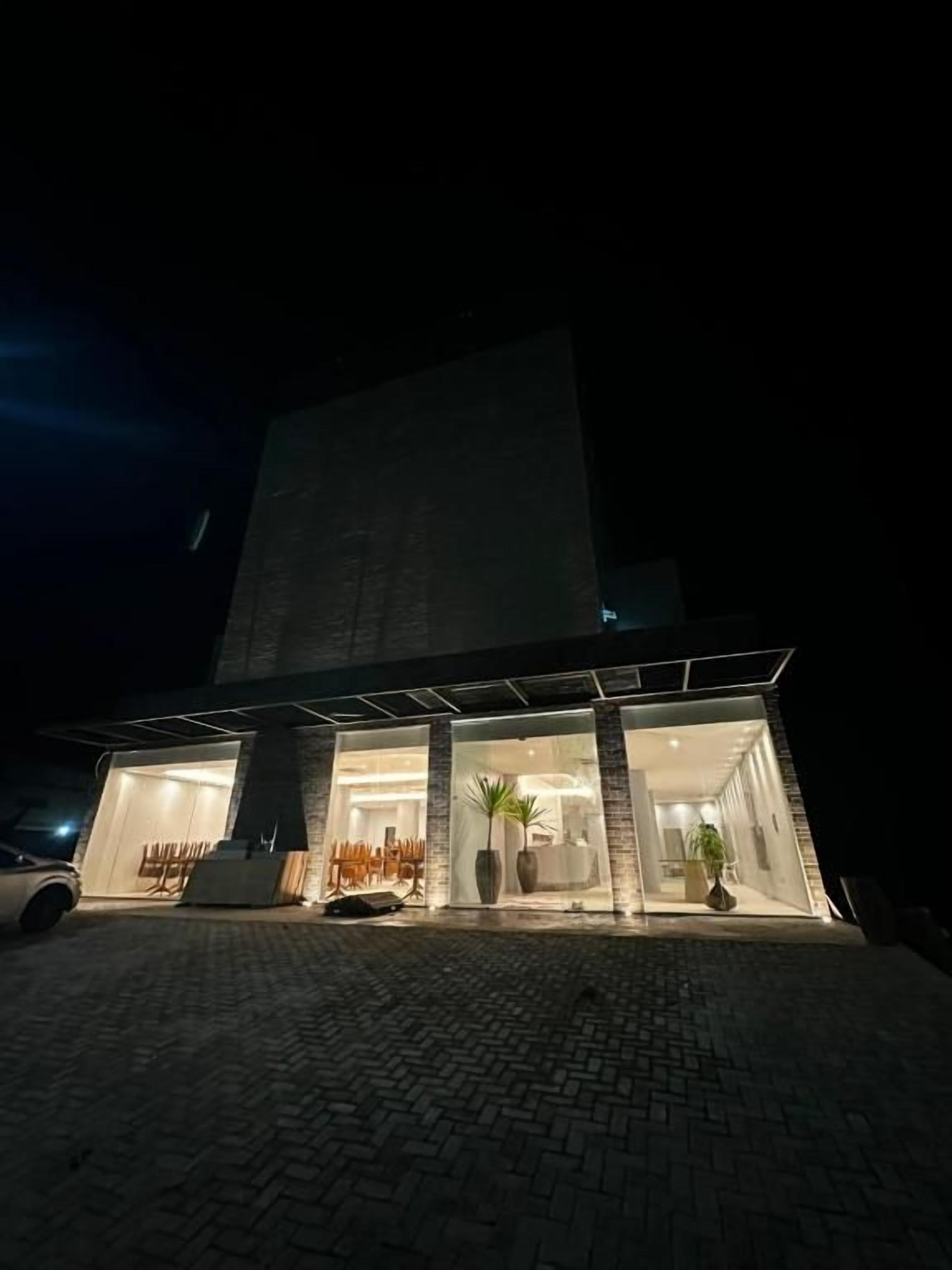 Um edifício moderno com fachada de grandes vidros, área de entrada pavimentada e um carro estacionado em frente.