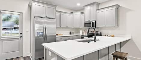 A modern kitchen with white cabinetry, stainless steel appliances, a large island with a sink, and a window with blinds.