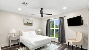 Una camera da letto con un letto, comodini, una sedia, una ventola a soffitto, una televisione e una finestra con tende.