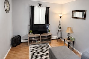 A living room with a flat-screen TV on a wooden stand, a floor lamp, a small table with a lamp, a star decoration on the wall, and a black fan. - Graceful Stays: Cozy 1BR in the Highlands (Louisville)